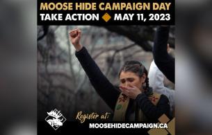Moose hide campaign supporter standing with one hand over her mouth while the other is a raised fist in the air at a campaign event