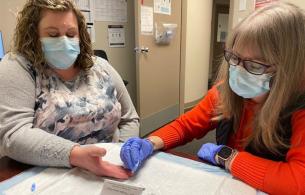 Two people wearing medical masks perform a blood spot test.