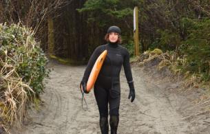  Dr. Jocelyn Black, head to toe in a black wet suit, walking down a beach pathway toward the water, holding her surf bard under her right arm.