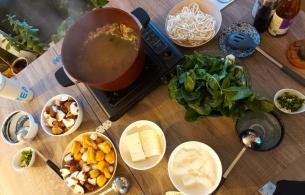 Several bowls containing noodles, tofu, greens, sauces and a large pot sitting on a table