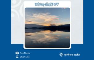 Calm waters reflecting clouds, shadows of mountains, and the orange setting sun of the background. 