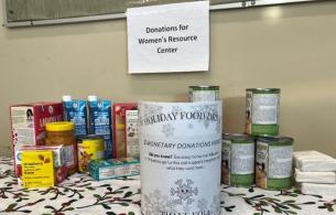 A stack of non-perishable food cans sits on a table covered with a holiday theme table cloth