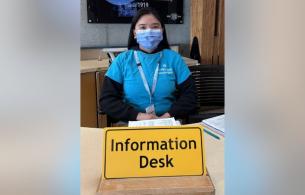 Diane sitting behind a yellow sign that reads "Information Desk"