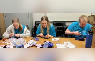 Volunteers knitting purple hats with supplies scattered across the table.