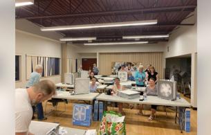 Participants sit at long tables with partially constructed DIY air cleaners standing next to them.