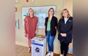 Three women stand beside a car seat box in a hallway in front of the Spirit of the North Healthcare Foundation sign.