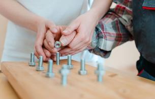 Two hands over a woodedn board attempting to screw nuts onto protruding bolts.