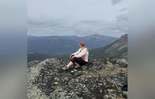 Amanda sits atop a mountain, viewing forests and mountains in the background. 