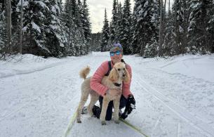 Haylee holding her dog on one knee in the snow, surrounded by trees